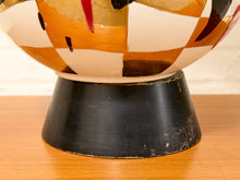 Load image into Gallery viewer, A close-up of a decorative ceramic bowl or vase with a black pedestal base, featuring a cream, orange, and brown geometric pattern with small red and black accents, resting on a wooden surface.

