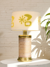 Load image into Gallery viewer, Vintage table lamp with textured cylindrical base, polished brass-tone accents, and off-white drum shade with yellow atomic starburst and abstract motifs
