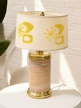 Load image into Gallery viewer, A mid-century modern table lamp with a cream-colored drum shade featuring yellow retro starburst and square motifs, a gold-finished metal top and base, and a cylindrical body wrapped in horizontal beige and pink stripes, set on a white surface against a white brick wall.
