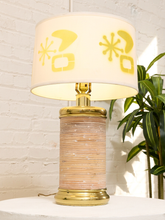 Load image into Gallery viewer, A vintage-style table lamp with a cream-colored drum shade decorated with yellow mid-century abstract motifs, a gold-finished metal base and collar, and a textured beige and gold striped cylindrical body, set against a white brick wall with a green plant in the background.
