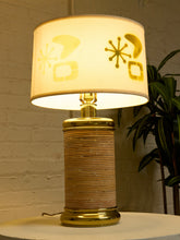 Load image into Gallery viewer, A vintage-style table lamp with a warm glowing cream-colored drum shade featuring yellow retro geometric cutouts, set on a light-colored surface against a white brick wall with a green plant in the background.
