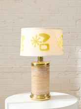 Load image into Gallery viewer, A table lamp with a white drum shade decorated with yellow retro mid-century modern geometric and starburst patterns, set on a white cylindrical pedestal against a white brick wall background.
