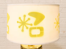 Load image into Gallery viewer, A white drum lampshade with a yellow mid-century modern geometric pattern, shown on a table lamp with a gold base against a white brick wall background.
