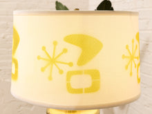 Load image into Gallery viewer, A close-up of a cylindrical lampshade with a warm cream fabric body and a yellow mid-century modern stencil pattern, set against a white brick wall background.
