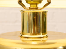 Load image into Gallery viewer, Close-up of a polished brass lamp base with a cylindrical column and curved top detail, reflecting warm golden light against a softly blurred light background.
