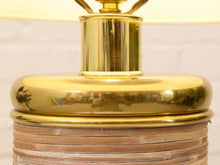 Load image into Gallery viewer, Close-up of a brass table lamp base with a polished gold-toned metal collar and a textured, ribbed brown base, set against a light brick wall background.
