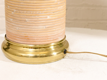 Load image into Gallery viewer, Close-up of a table lamp with a ribbed beige shade and a polished gold base, sitting on a white fabric surface with a light brick wall in the background.
