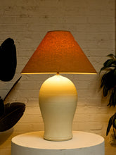 Load image into Gallery viewer, A table lamp with a textured orange-brown conical shade and a glossy cream-to-ivory gradient ceramic base, illuminated against a white brick wall backdrop.
