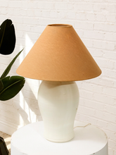 Load image into Gallery viewer, A table lamp with a tapered tan paper shade and a glossy white ceramic base, displayed on a white pedestal against a white brick wall with a green plant leaf on the left.
