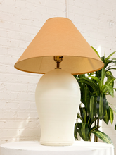 Load image into Gallery viewer, A table lamp with a warm beige fabric drum shade and a cream-colored ceramic base, displayed on a white table against a white brick wall with a green plant in the background.
