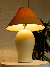 Load image into Gallery viewer, A table lamp with a warm brown fabric drum shade and a cream-colored ceramic base, illuminated to cast a yellow glow, set against a white brick wall with a green plant in the background.
