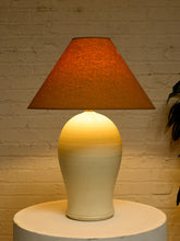 Load image into Gallery viewer, A table lamp with a textured orange-brown conical shade and a glossy cream-to-ivory gradient ceramic base, illuminated against a white brick wall backdrop.
