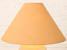 Load image into Gallery viewer, A warm beige fabric lampshade glowing with a soft yellow light, set against a white painted brick wall background.
