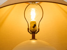Load image into Gallery viewer, Close-up of a lit vintage-style bulb in a brass-colored metal fixture, set against a warm yellow fabric shade.
