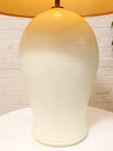 Load image into Gallery viewer, A ceramic table lamp with a rounded, bulb-shaped off-white base and a warm yellow-orange fabric shade, set against a white brick wall background.
