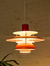 Load image into Gallery viewer, A modern multi-tiered pendant light with a red top cap and orange lower shade, glowing with warm light against a textured beige brick wall.
