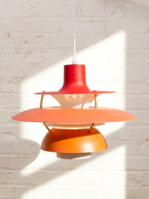 Load image into Gallery viewer, A mid-century modern multi-tier pendant lamp with red, orange, and peach shades, suspended by a white cord against a white brick wall with strong sunlight and shadows.
