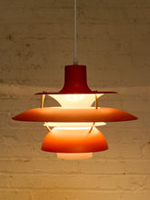 Load image into Gallery viewer, A mid-century modern hanging pendant lamp with a red metal top canopy, orange glass shades, and brass-colored support rods, suspended against a warm, off-white brick wall.
