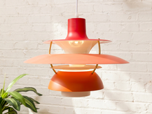 Load image into Gallery viewer, A mid-century modern multi-tier pendant lamp with red, orange, and peach-pink metal shades, suspended against a white brick wall with a green plant leaf visible in the lower left corner.
