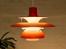 Load image into Gallery viewer, A mid-century modern tiered pendant light with red, orange, and white shades, shown illuminated against a warm brick wall background.
