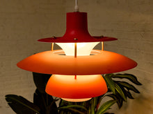 Load image into Gallery viewer, A mid-century modern hanging pendant lamp with three concentric shades in red, orange, and white, glowing warmly against a light brick wall with green leaves in the foreground.
