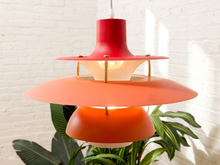 Load image into Gallery viewer, A mid-century modern tiered pendant light with red, orange, and coral shades, shown hanging in front of a white brick wall with green plants.
