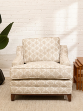 Load image into Gallery viewer, A single upholstered accent chair with a beige and off-white geometric lattice pattern, featuring dark brown wooden legs, set against a white brick wall background.
