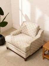 Load image into Gallery viewer, A beige armchair with a light beige and white geometric trellis pattern, styled in a bright room with a white brick wall, a potted plant, and a wooden side table.
