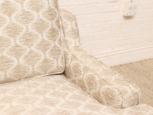 Load image into Gallery viewer, Close-up of an upholstered armchair corner with a beige and off-white geometric patterned fabric, set against a white brick wall and carpet.
