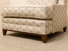 Load image into Gallery viewer, A close-up of a upholstered armchair or club chair showing the front cushion, side panel, and wooden legs. The fabric has a warm beige and off-white geometric trellis pattern, and the chair sits on a textured beige rug.
