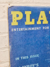Load image into Gallery viewer, A vintage blue Playboy magazine cover mounted against a white brick wall. The cover features the iconic yellow 'PLAYBOY' logo at the top, with white text reading 'ENTERTAINMENT FOR' and 'IN THIS ISSUE' below.
