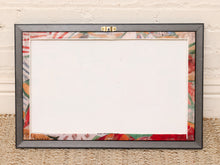 Load image into Gallery viewer, A rectangular picture frame with a dark gray outer border and a colorful patterned inner border, shown standing against a white brick wall on a woven rug.
