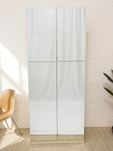 Load image into Gallery viewer, A tall, narrow white storage cabinet or wardrobe with four front doors, shown standing against a white brick wall in a styled interior. A tan chair is partially visible on the left, and a plant leaf appears on the right.
