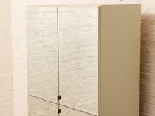 Load image into Gallery viewer, A modern wall-mounted bathroom cabinet with two mirrored doors and a solid beige side panel, set against a white brick wall.
