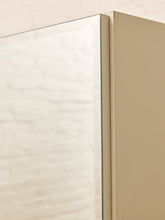 Load image into Gallery viewer, Close-up of a beige medicine cabinet door with a clear glass front, showing the top edge and a soft reflection of a bathroom wall.
