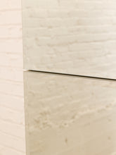 Load image into Gallery viewer, A close-up of a modern wall mirror with a thin black frame, reflecting a warm off-white brick wall.
