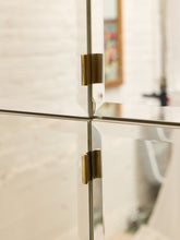 Load image into Gallery viewer, Close-up of a modern glass cabinet or door with brass hinges, showing the reflective surfaces and hardware against a softly blurred interior background.
