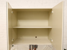 Load image into Gallery viewer, An open beige storage cabinet with two doors swung open, showing two interior shelves. The cabinet is mounted against a white brick wall, with a white drop cloth covering the floor and a person's legs visible at the bottom edge.
