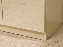 Load image into Gallery viewer, Close-up of the corner of a light beige or cream cabinet with mirrored doors, resting on a textured beige woven rug.
