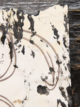 Load image into Gallery viewer, A close-up of a distressed off-white surface with black splattered marks and curved brownish lines, bordered on the right by a dark textured vertical element.
