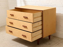 Load image into Gallery viewer, A mid-century modern wooden nightstand with three drawers pulled out, featuring a light natural wood finish, dark brown drawer pulls, and tapered dark legs, set against a white brick wall and beige carpet.
