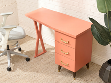 Load image into Gallery viewer, A coral pink three-drawer desk with gold handles and dark legs, styled in a home office next to a white office chair and a potted plant.

