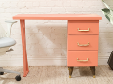 Load image into Gallery viewer, A coral pink mid-century modern writing desk with three drawers on the right side, featuring brass handles and tapered wooden legs with gold caps, set against a white brick wall.
