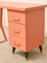 Load image into Gallery viewer, A mid-century modern three-drawer nightstand in a warm coral pink finish, featuring brass handles and tapered dark wood legs with gold caps, set against a white brick wall and beige carpet.
