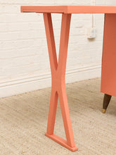 Load image into Gallery viewer, A close-up of a mid-century modern table leg in a warm coral or salmon pink finish, standing on a beige textured rug against a white brick wall.
