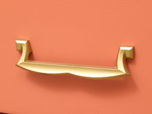Load image into Gallery viewer, A modern curved brass drawer pull handle mounted on a textured coral-pink drawer front.
