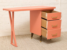 Load image into Gallery viewer, A mid-century modern writing desk with a coral pink finish, featuring three open drawers with brass handles and wooden legs, set against a white brick wall and beige carpet.
