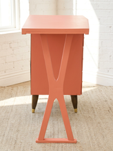 Load image into Gallery viewer, A mid-century modern side table with a terracotta orange-pink painted finish, featuring a geometric X-shaped pedestal base and dark wood legs with brass-colored feet, set in a bright room with a white brick wall and woven rug.

