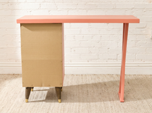 Load image into Gallery viewer, A mid-century modern desk with a coral pink painted top and legs, paired with a natural cardboard side cabinet and light wood feet, set against a white brick wall and beige rug.
