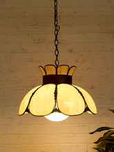 Load image into Gallery viewer, A vintage-style pendant light with a warm yellow glass shade shaped like layered petals, suspended by a dark metal chain against a white brick wall.
